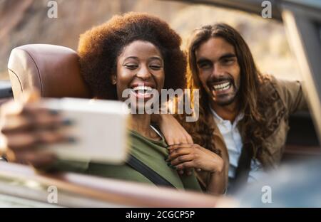 Happy young couple taking selfie with mobile smartphone while doing road trip - Travel people having fun driving in convertible car Stock Photo