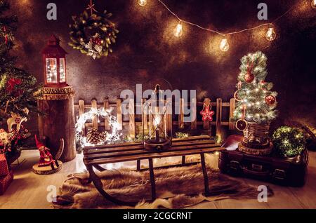 Christmas theme arrangement in dark evening home room. Lot of party string lights illuminated, lanterns, old retro sleight, tiny Christmas tree. Stock Photo