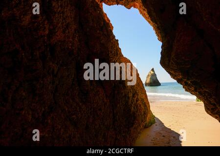 A stunning view from inside a small cave, looking out onto the beach in Algarve, Portugal. In the distance, a triangular rock formation. Stock Photo