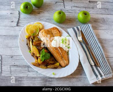 wild salmon fillet with roated potatoes and tatar sauce served on a white plate with knife and fork on a wooden light table Stock Photo