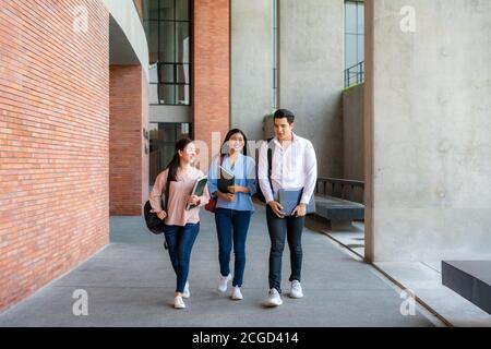 Asian three students are walking and talking together in university hall during break in University. Education, Learning, Student, Campus, University, Stock Photo