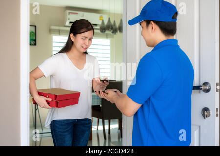 Asian delivery young man in blue uniform smile and holding food boxes in front house and Asian woman accepting a delivery of food boxes and payment QR Stock Photo