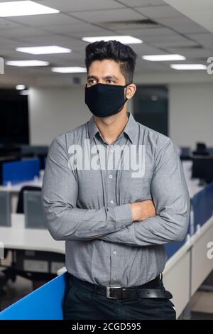 Portrait of handsome young Indian businessman wearing Covid protection mask standing in office, corporate environment, standing with arms crossed. Stock Photo