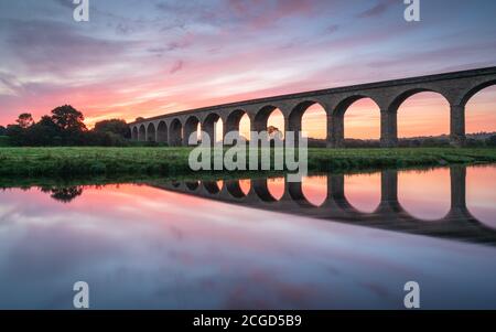 Arthington Viaduct is reflected in the River Wharfe during a colourful sunrise, the vibrant coulours in the sky mirrored by the water. Stock Photo