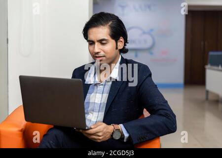 Portrait of young successful Indian business man in dark suit sitting on couch and working on laptop, Corporate environment. Stock Photo