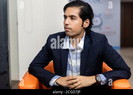 Portrait of young successful Indian business man in dark suit sitting on couch in a corporate environment, looking away from camera. Stock Photo
