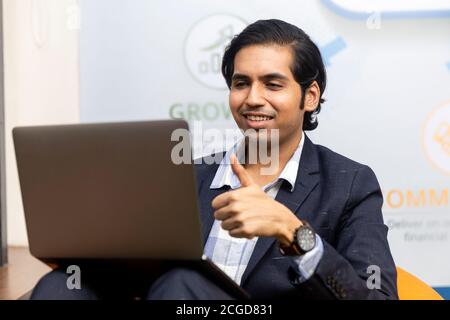 Portrait of young successful Indian business man in dark suit sitting and working on laptop, Corporate environment, expressive face. Stock Photo