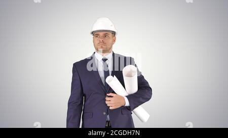 Male architect in a suit and hard hat walking with blueprints and briefcase on gradient background. Stock Photo