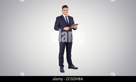 Expressive young businessman with tablet presenting something swiping on gradient background. Stock Photo