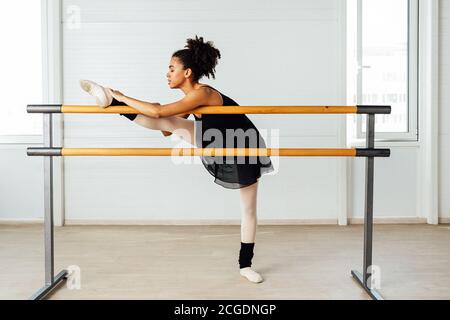 Female ballet dancer with black hair stretching her leg. Side view of young woman preparing for a ballet class in studio Stock Photo