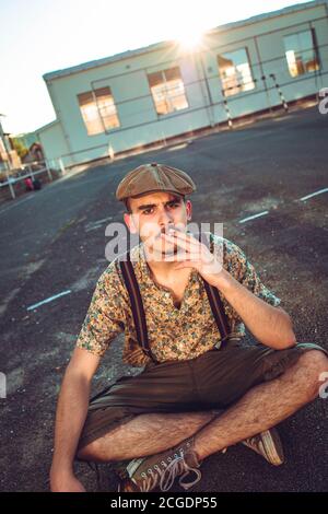 Young hipster man in retro clothing sits on the ground and smokes Stock Photo