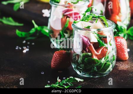 Fresh salad with spicy arugula, goat cheese, strawberry and prosciutto or ham in modern glass jars on black table, selective focus Stock Photo