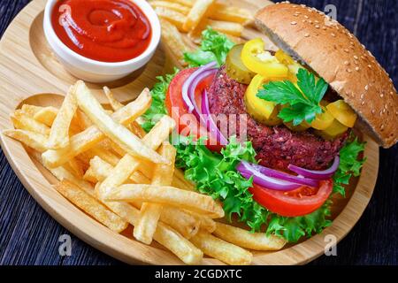 Plant-based vegan beet burger with pickles fresh lettuce, tomatoes, red onions inside of sesame seed bun french fry, ketchup on a bamboo plate on a da Stock Photo