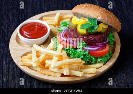 Vegan beet burger with pickles fresh lettuce, tomatoes, red onions, french fry, ketchup on a bamboo plate on a dark wooden table, top view, close-up Stock Photo