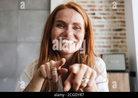 Image of happy ginger woman with freckles smiling and pointing fingers at camera in home Stock Photo