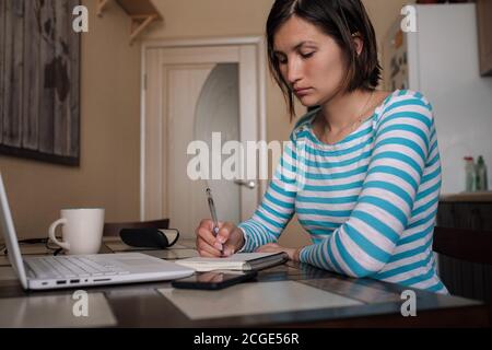 Serious beautiful young adult asian woman taking notes while sitting in front of laptop computer on kitchen table near window. Female student involved Stock Photo
