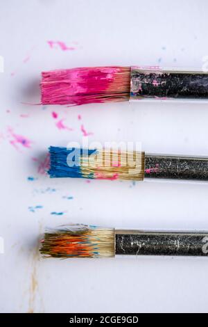 Three small paint brushes covered in different coloured paint on a white background Stock Photo
