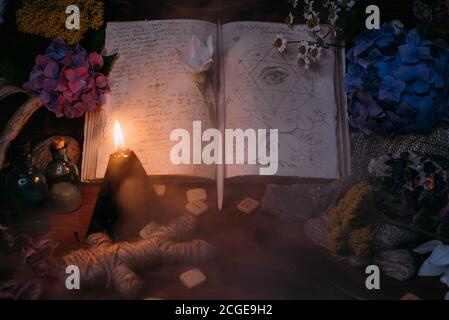 Open old book with magic spells, runes, black candle and herbs on witch table. Occult, esoteric, divination and wicca concept. Halloween vintage backg Stock Photo