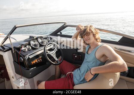 Young and handsome guy of european appearance wearing sunglasses and colourful beach clothes, sitting at the helm, driving speedboat on a sunny day. Stock Photo