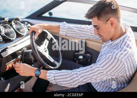 Serious handsome business man dressed in classy style, with sunglasses relaxing during sailing on river, he sitting begins the helm and driving it on Stock Photo