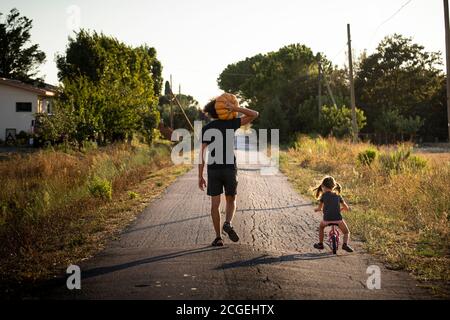 Little child girl, riding a bike, with her young father carrying a big halloween pumpkin on a country road at sunset. Back view. Stock Photo