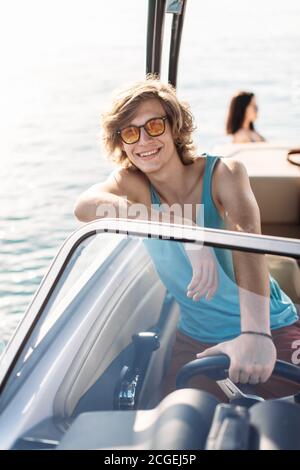 Young and handsome guy of european appearance wearing sunglasses and colourful beach clothes, sitting at the helm, driving speedboat on a sunny day. Stock Photo