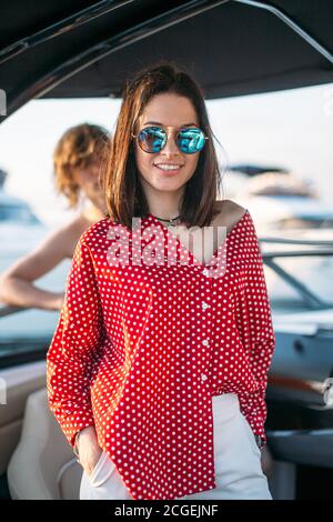 Outdoor portrait of cute brunette lady, fashionably dressed in red blouse, wearing trendy sunglasses, smiling at camera, in anticipation of intresting