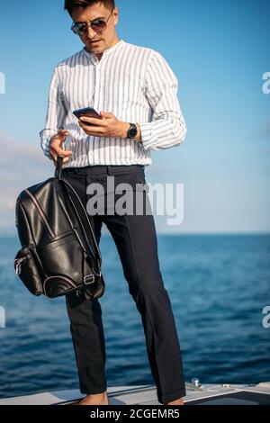 Confident caucasian business man with lether backpack handling an order over the phone and with his laptop while sailing on yacht trip at sea Stock Photo