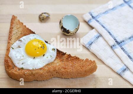 Toasted bread with fried quail egg on a wooden cutting board with a kitchen towel and eggshells, selective focus. Healthy breakfast. Stock Photo