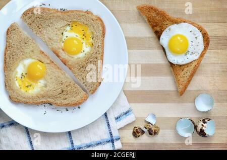Fried bread slices with quail eggs on a white plate placed on a wooden cutting board with a kitchen towel and eggshells, top view. Stock Photo