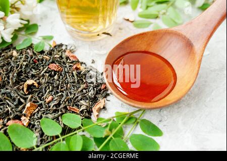 wooden spoon with honey, dried tea leaves with berries and acacia flowers broth in glassware cup with lush flowering white acacia tree Branch, useful Stock Photo