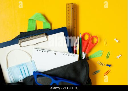 black open backpack filled with school accessories and teaching supplies and a protective mask on a bright yellow background. Returning to school in a Stock Photo