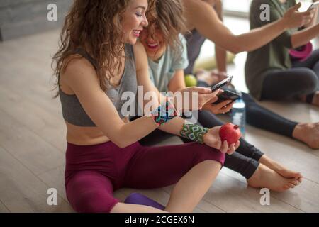 Fitness friends, man and three woman sitting on floor, having light fruit snack and watching photos on smartphone after successful yoga training in gy Stock Photo