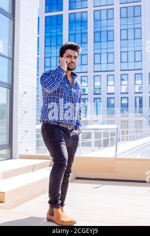 Indian bearded owner of company in casual wear having friendly conversation on modern smartphone standing against city office buildings Stock Photo