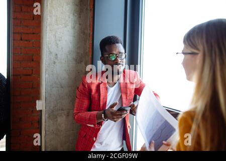 Multiethnic group of students chilling at university hall during recess, standing by the window talking or communicating in social media via laptop Stock Photo