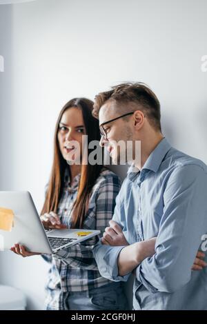 Two heterosexual architects collegues discussing project of new contemporary skyscraper using laptop, standing against white office wall Stock Photo