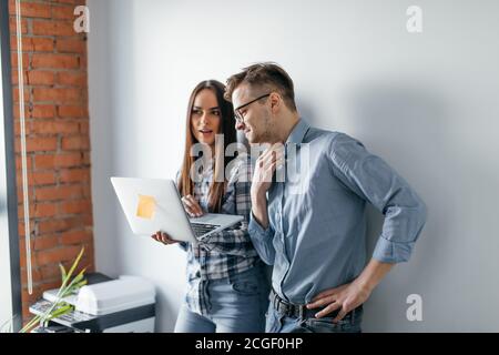 Two heterosexual architects collegues discussing project of new contemporary skyscraper using laptop, standing against white office wall Stock Photo