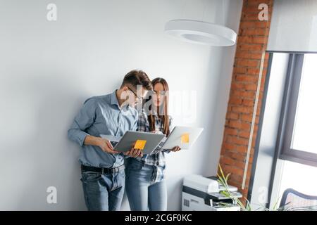 Two heterosexual architects collegues discussing project of new contemporary skyscraper using laptop, standing against white office wall Stock Photo