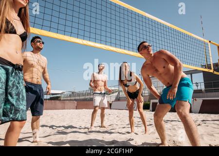 Young positive caucasian people in good physical form playing volleyball on beach. Athletic men and women enjoying active leisure time outdoors on vac Stock Photo