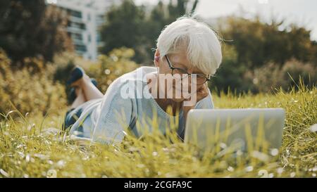 senior retired woman using laptop in the nature while lying on the grass. High quality photo Stock Photo