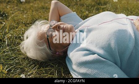 senior retired woman with headphones lying on the grass and listening to the music. High quality photo Stock Photo