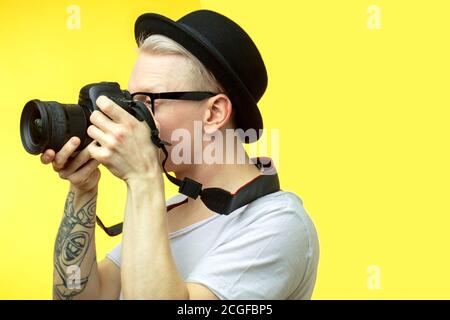 Half-length portrait of young handsome albino man tourist wearing black hat with retro camera in hands isolated over yellow studio wall Stock Photo