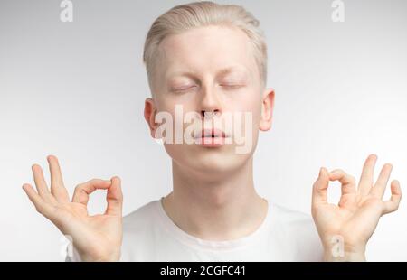 Serene albino man holding fingers in mudra, with eyes closed, practicing group meditation in studio tries to relax, poses against white background. Pe Stock Photo
