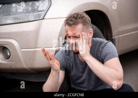 Young man sits with bloodied head near wheel of car, screaming and regretting act Stock Photo