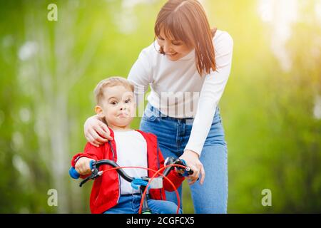 Mom teaches boy child to ride bicycle, balance holding training. In background is sunny day park Stock Photo