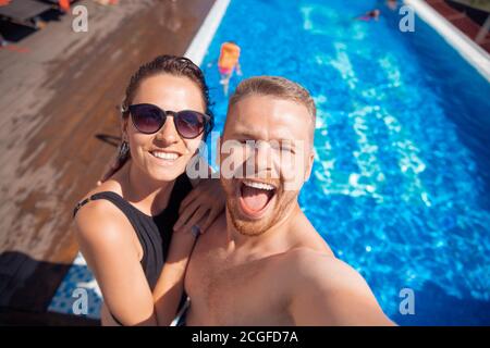 Couple of friends make selfie photo, happy caucasian beautiful young woman and man hug traveling on background hotel pool Stock Photo