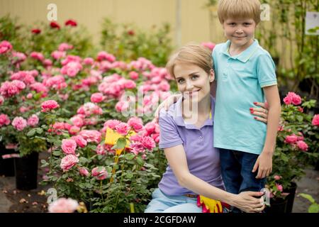 Young attractive mother hugging her 5-6 year old sons miling at camera near roses in a summer garden Stock Photo