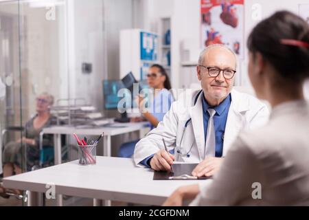 Senior doctor talking with patient about her illness in hospital office. Nurse holding x-ray image in the background. Elderly aged woman in wheelchair. Stock Photo