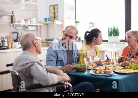 Son looking at disabled senior father in wheelchair during family lunch with healthy food. Happy Mature parents. Stock Photo