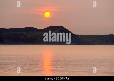 Lyme Regis, Dorset, UK.  11th September 2020.  UK Weather.  View from Lyme Regis in Dorset at sunrise as the sun rises up through the thin hazy cloud from behind Golden Cap on the Jurassic Coast.  Picture Credit: Graham Hunt/Alamy Live News Stock Photo